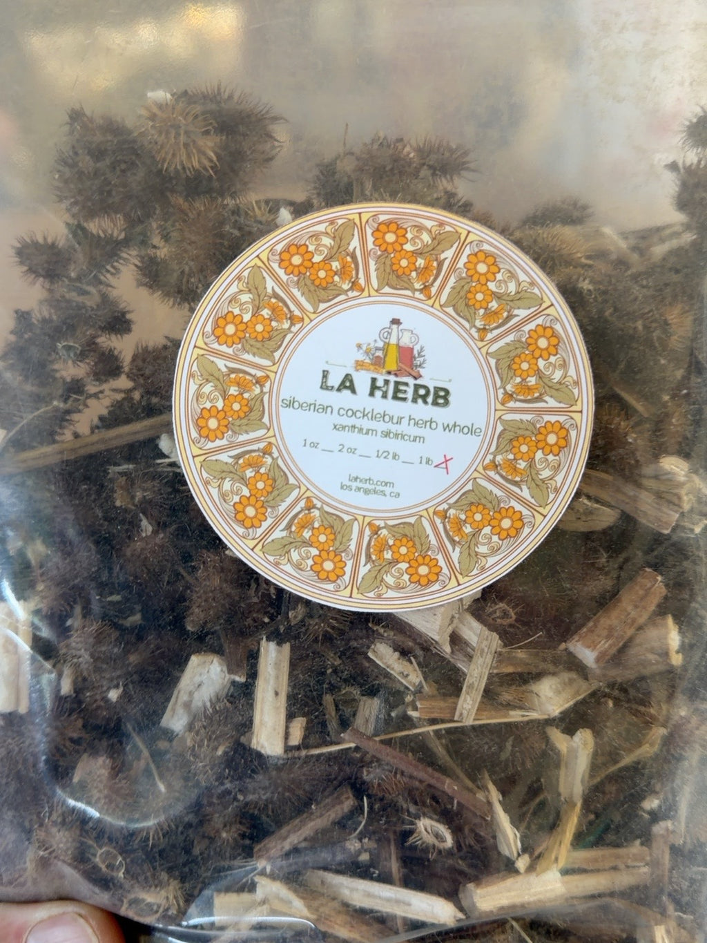 Original LA Herb supplier package labeled Siberian cocklebur herb whole, Xanthium sibiricum, showing dried burs and stems.